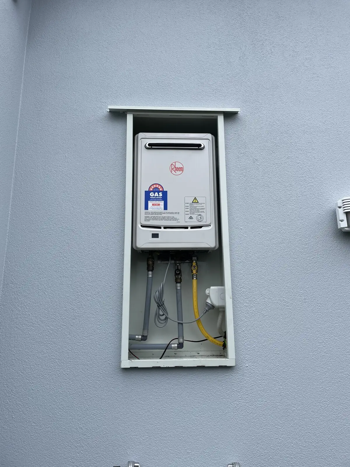 Rheem continuous flow gas water heater with its gas energy rating label, mounted in a weather box on a grey exterior wall