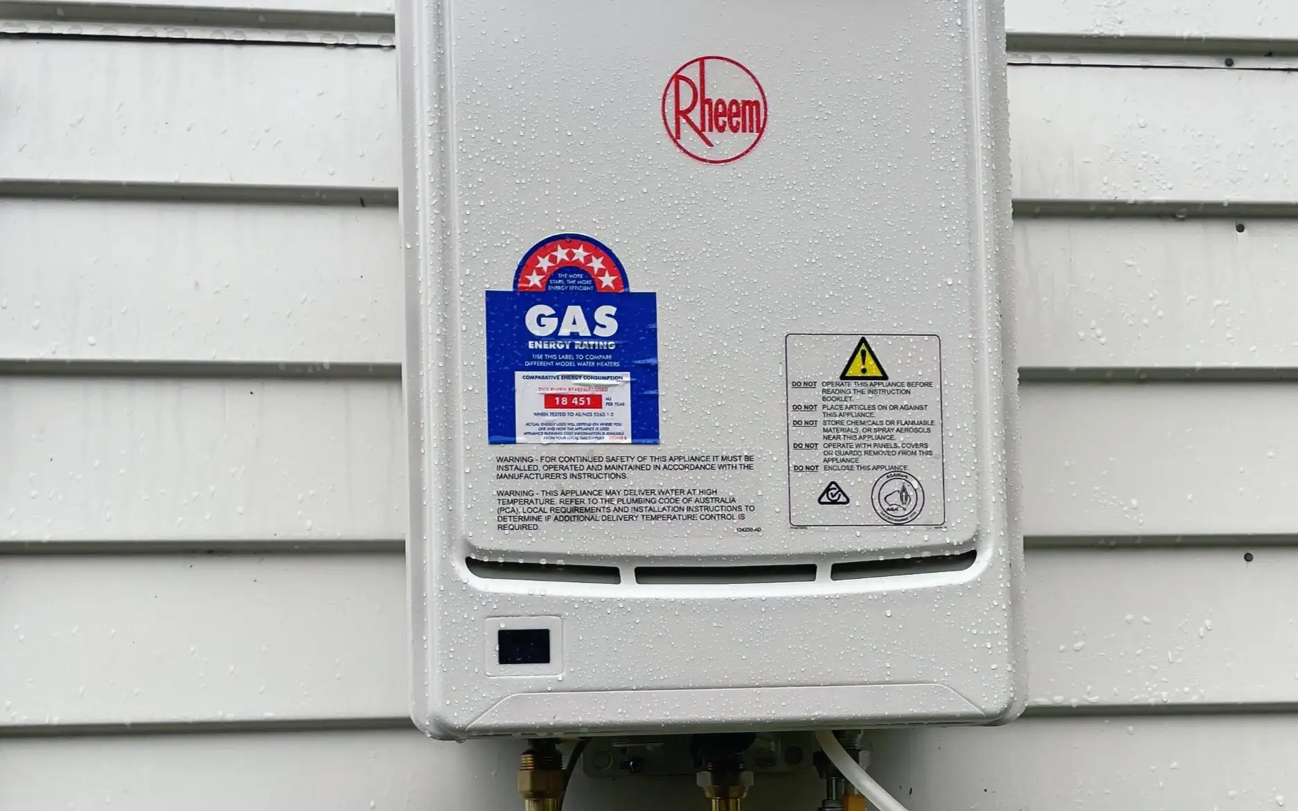 Rheem continuous flow gas water heater on a weatherboard wall, with a digital gas pressure gauge connected below it during commissioning