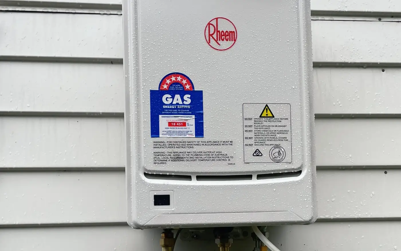 Rheem continuous flow gas water heater on a weatherboard wall, with a digital gas pressure gauge connected below it during commissioning