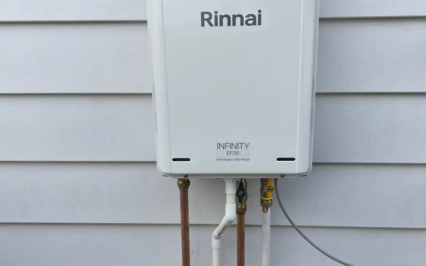 Rinnai INFINITY gas continuous flow water heater mounted on a weatherboard wall, with its pipework and outdoor power point below